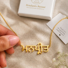 Charger l'image dans la galerie, A gold necklace with a custom Devanagari script pendant held by a hand, with a white jewelry box and thank you card in the background on a beige fabric surface.