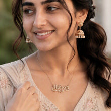 Load image into Gallery viewer, A woman with dark hair styled in a half-updo, wearing a beige embroidered traditional outfit, a gold nameplate necklace, and jhumka earrings, set against a softly blurred outdoor background.
