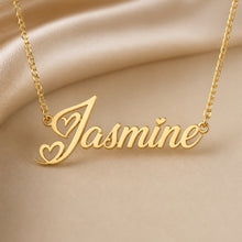 Load image into Gallery viewer, A gold nameplate necklace spelling 'Jasmine' with heart details, displayed on a draped beige satin fabric background.

