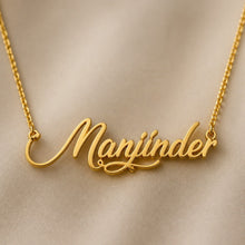 Load image into Gallery viewer, A gold nameplate necklace spelling 'Manjinder' in elegant cursive script, displayed on a soft beige fabric background.
