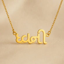 Load image into Gallery viewer, A gold nameplate necklace featuring stylized Arabic calligraphy, suspended from a delicate gold chain against a warm beige background.

