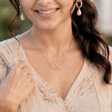 Load image into Gallery viewer, A woman with dark hair tied back wears a light beige embroidered traditional outfit and gold jewelry, including a necklace with the Hindi name 'Anurag' and matching earrings, set against a softly blurred outdoor background.
